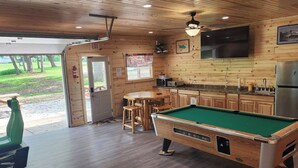 Une table de billard se trouve dans une pièce au plancher en bois, qui comprend également un coin cuisine, une table à manger et une porte donnant sur l’extérieur.