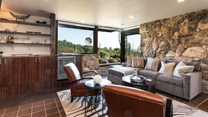 A modern living room with a stone wall, a grey sofa, leather chairs, and a kitchen area with wooden cabinets and a built-in oven. - 7 BD - 2 homes - Pool, Spa, Minutes to Plaza (Sonoma)