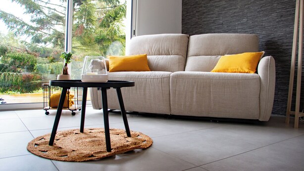 A modern living room with a sofa, two yellow cushions, a small table with a vase and a plant, and a view of trees through a large window. - New PMR-accessible gîte (Anould)