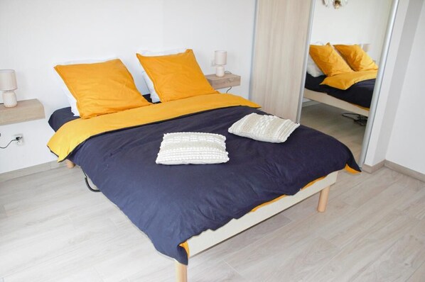A neatly made bed with a dark blue and yellow bedspread, two white pillows, and a white knitted blanket. There is a bedside table with a lamp and a small wooden nightstand with a lamp. A full-length mirror is visible on the wall. - New PMR-accessible vacation home (Anould)