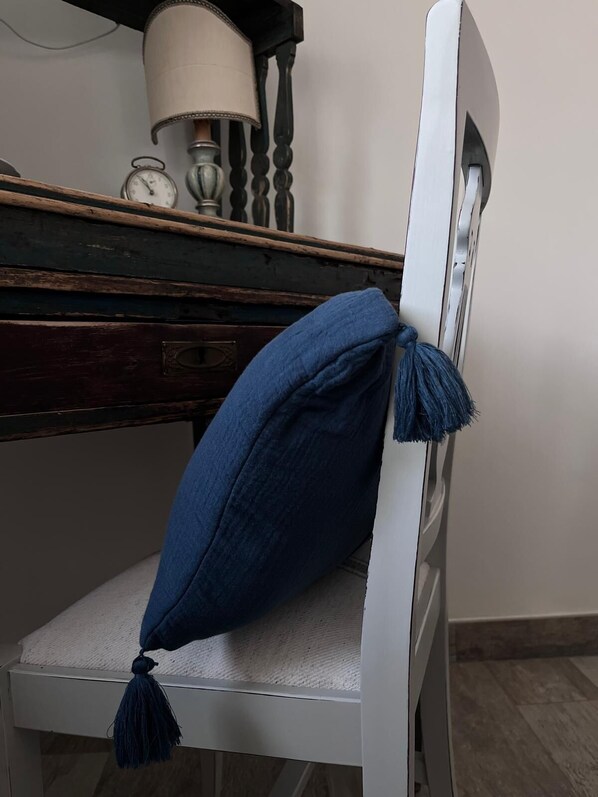 A white chair with a blue cushion and tassels, a wooden desk with a lamp, and a clock. - Clare's Garden_ Shelter for Weary Souls (Serrastretta)