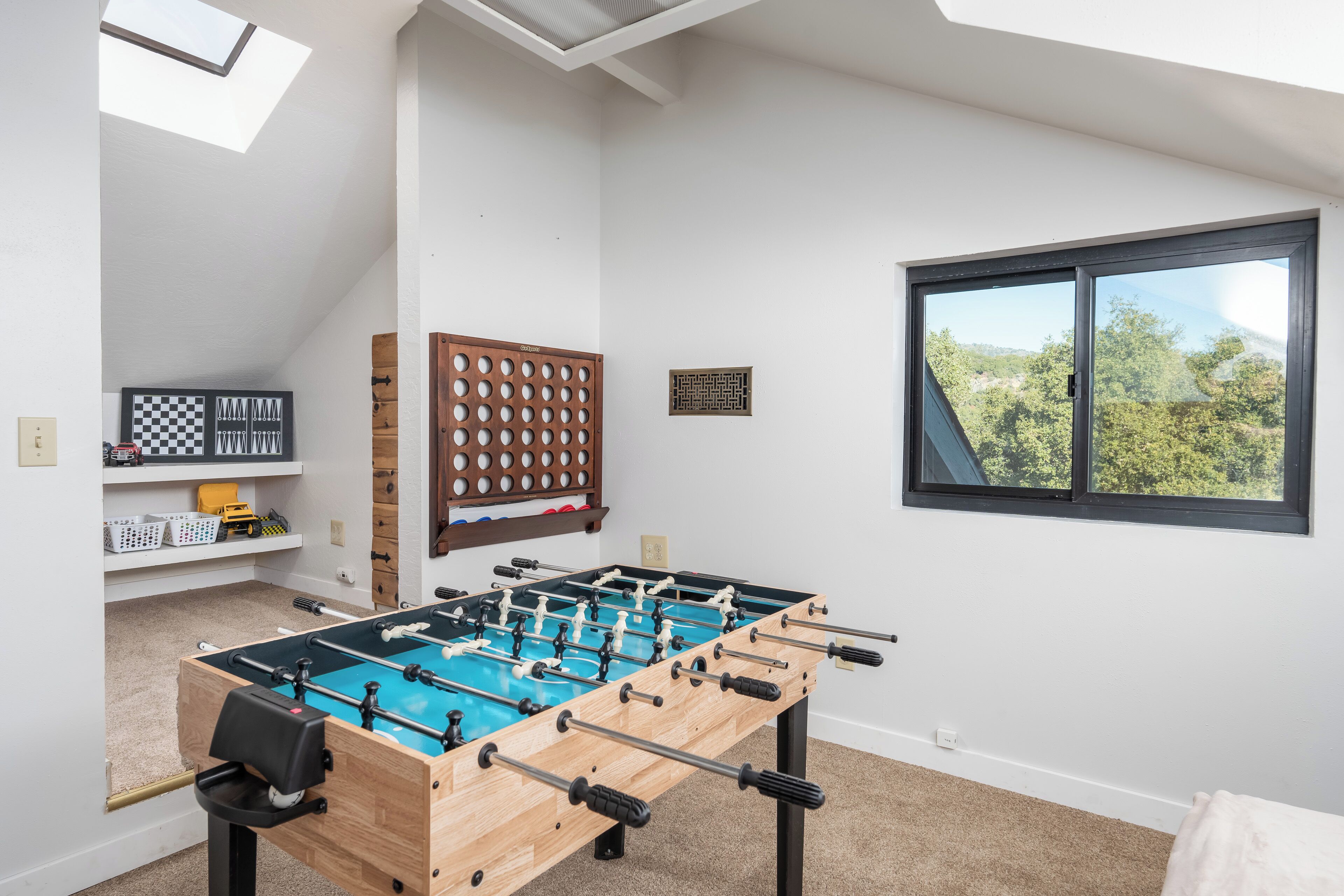 A room with a foosball table, a checkered board, and a window with a view of trees.
