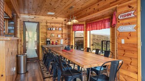 Interior de una cabaña de madera con una larga mesa de comedor, sillas negras, un cubo de basura y un letrero que dice 'Fishin' Cabin'.