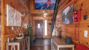 Interior de una cabaña de madera con una escalera de madera, una mesa de madera con un teléfono rojo, un mapa del mundo y un cuadro de un paisaje montañoso.