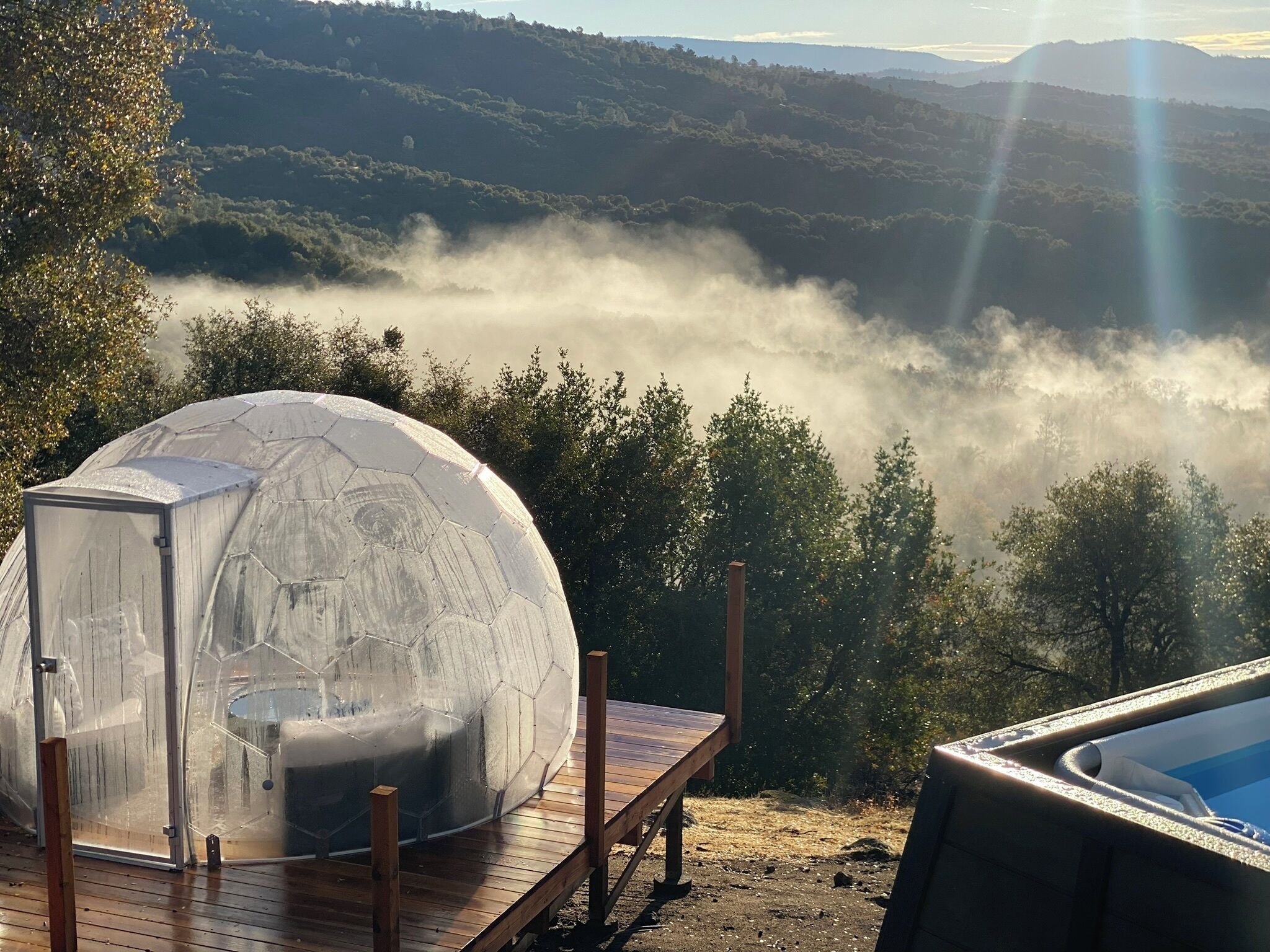 Una estructura de vidrio en forma de cúpula sobre una terraza de madera con vistas a un valle cubierto de niebla y colinas lejanas.
