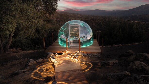 Una estructura abovedada transparente con una terraza de madera que la conecta, rodeada de rocas y árboles.