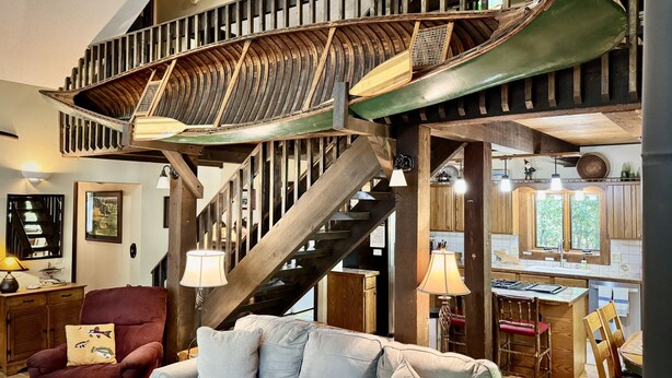 A spacious living area with a high wooden staircase, a comfortable sofa, and a dining area with a kitchen in the background.