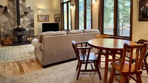A cozy living room with a stone fireplace, a sofa, a dining table with chairs, and large windows offering a view of trees.