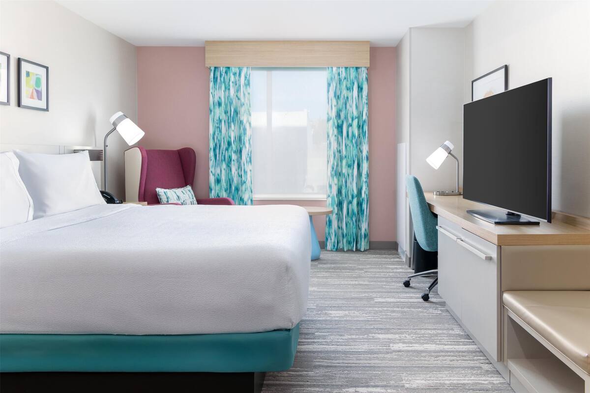 Habitación de hotel con cama, escritorio con televisor, silla, lámpara de escritorio y ventana con cortinas.