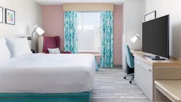 A hotel room with a bed, a desk with a television, a chair, a desk lamp, and a window with curtains.