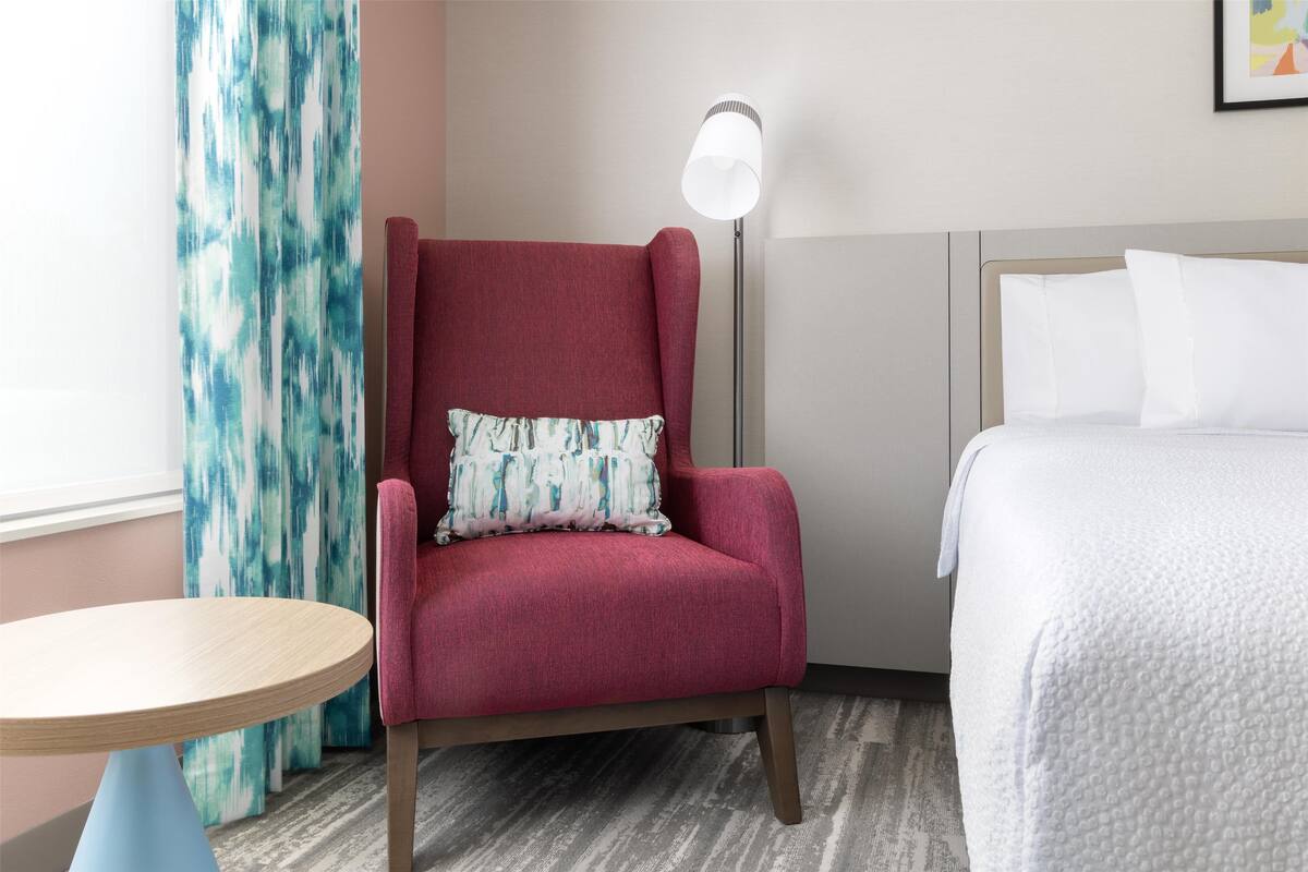 Habitación de hotel con un sillón rojo, una mesa redonda de madera, una cama con ropa blanca y una lámpara de pie.