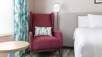 Habitación de hotel con un sillón rojo, una mesa redonda de madera, una cama con ropa blanca y una lámpara de pie.