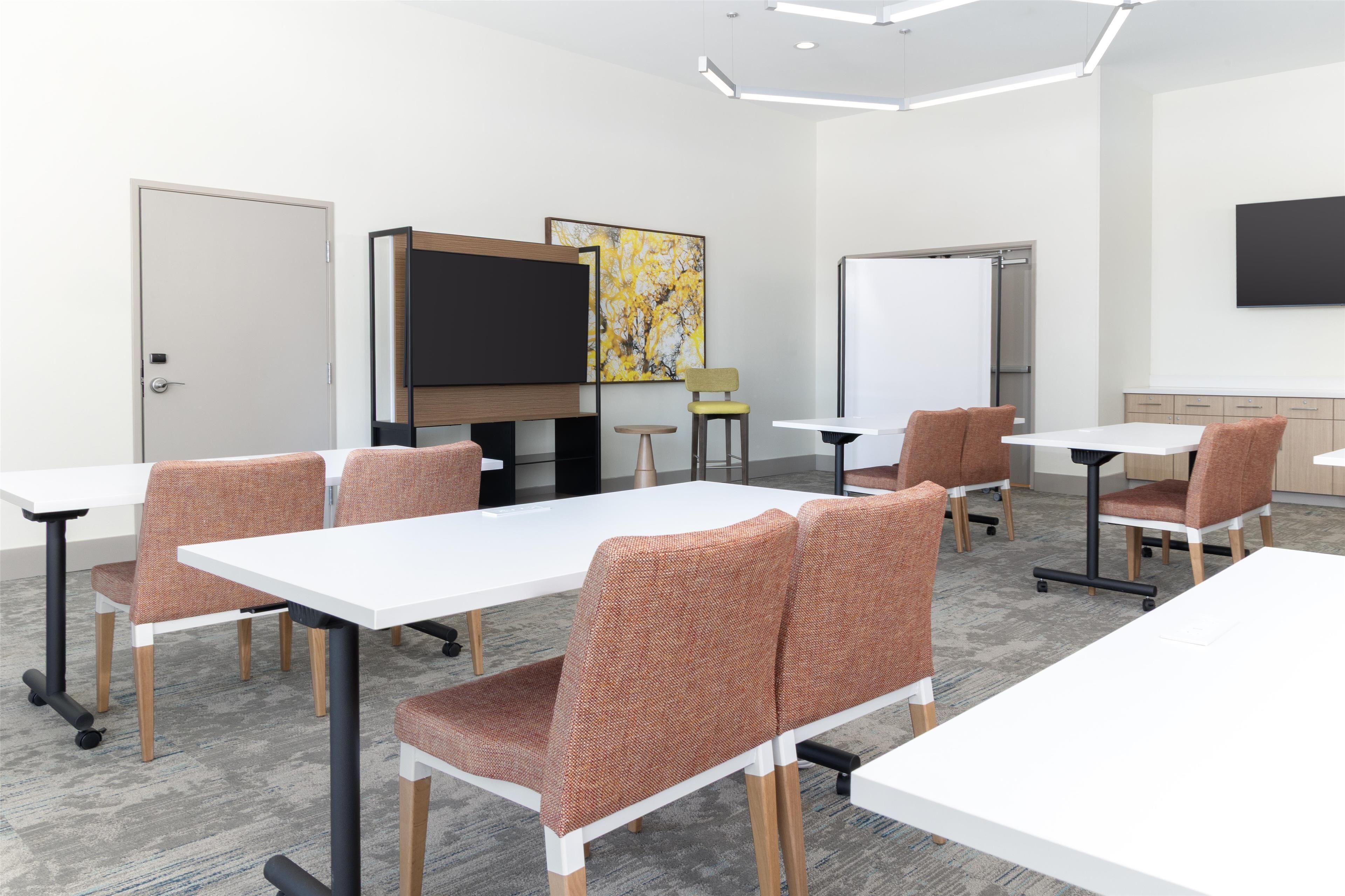 Une salle de conférence avec des tables blanches et des chaises marron, un tableau blanc et un grand tableau abstrait accroché au mur.