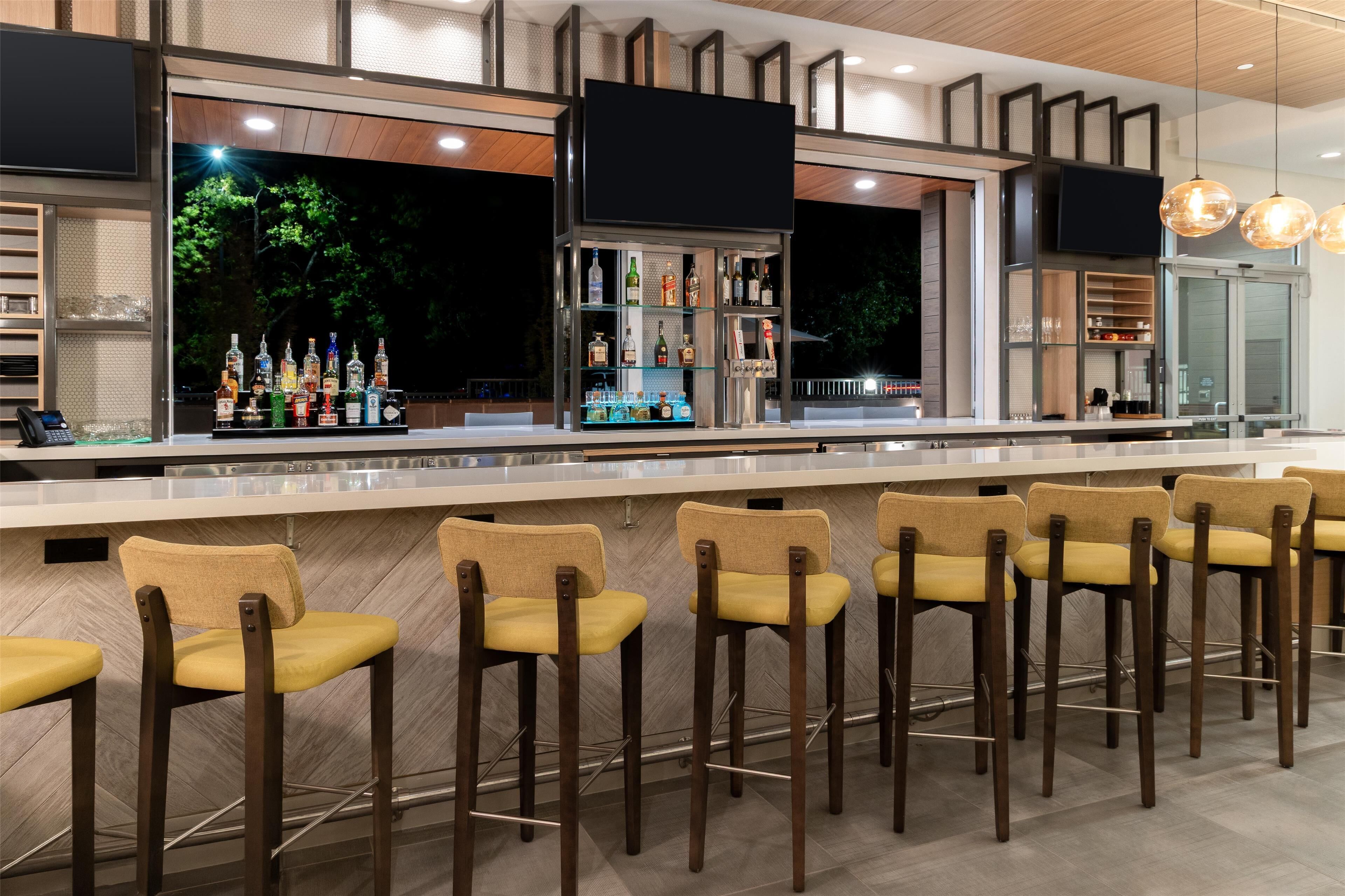 Zona de bar con varios taburetes, una barra y estantes llenos de botellas.
