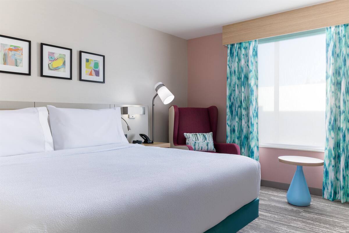 Una habitación de hotel con cama, escritorio con lámpara, silla y ventana con cortinas.