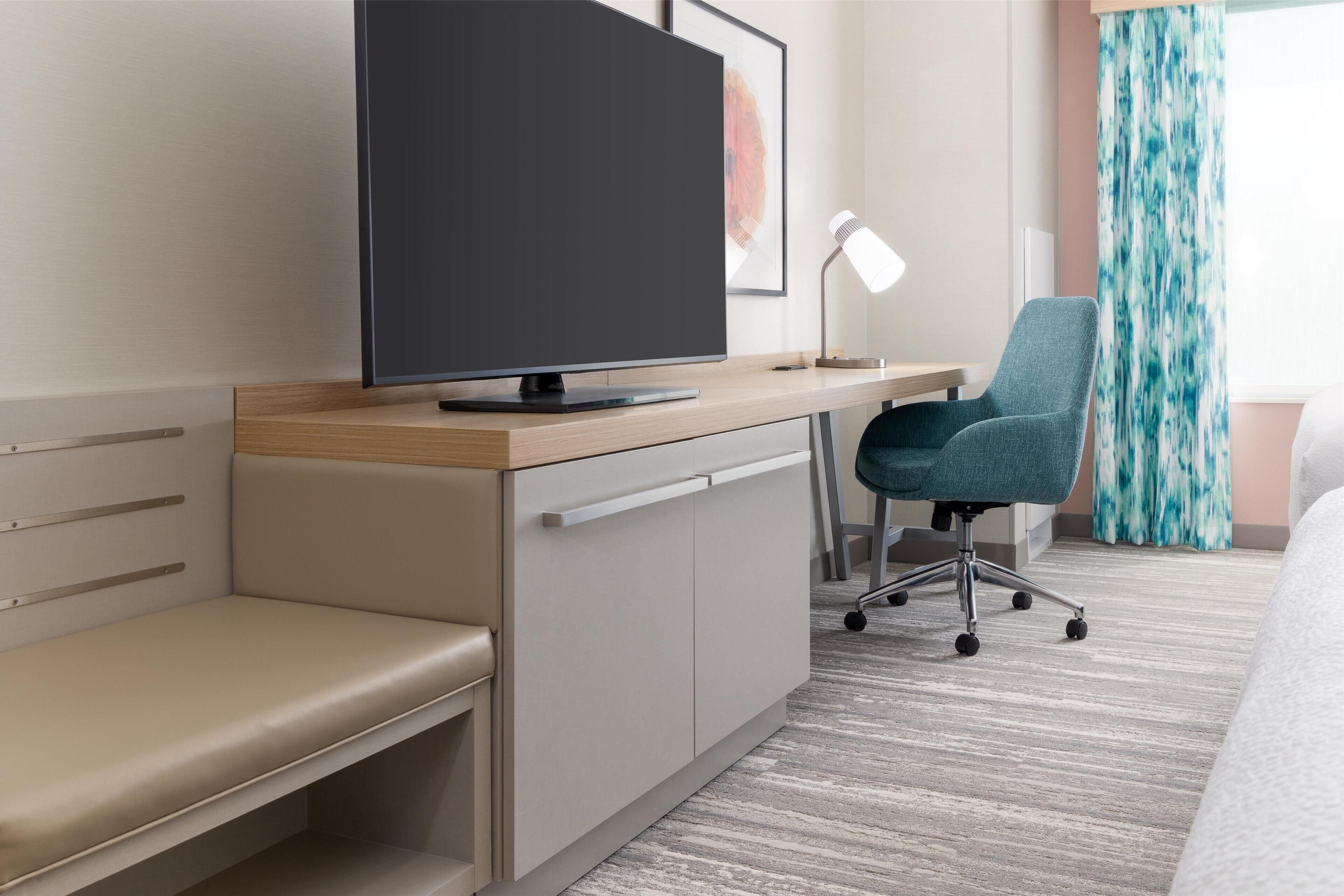 A modern hotel room with a wooden desk, a flat-screen TV, a blue office chair, a desk lamp, and a bench.
