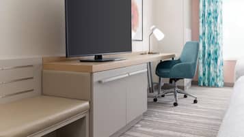 Una habitación de hotel moderna con un escritorio de madera, un televisor de pantalla plana, una silla de oficina azul, una lámpara de escritorio y un banco.
