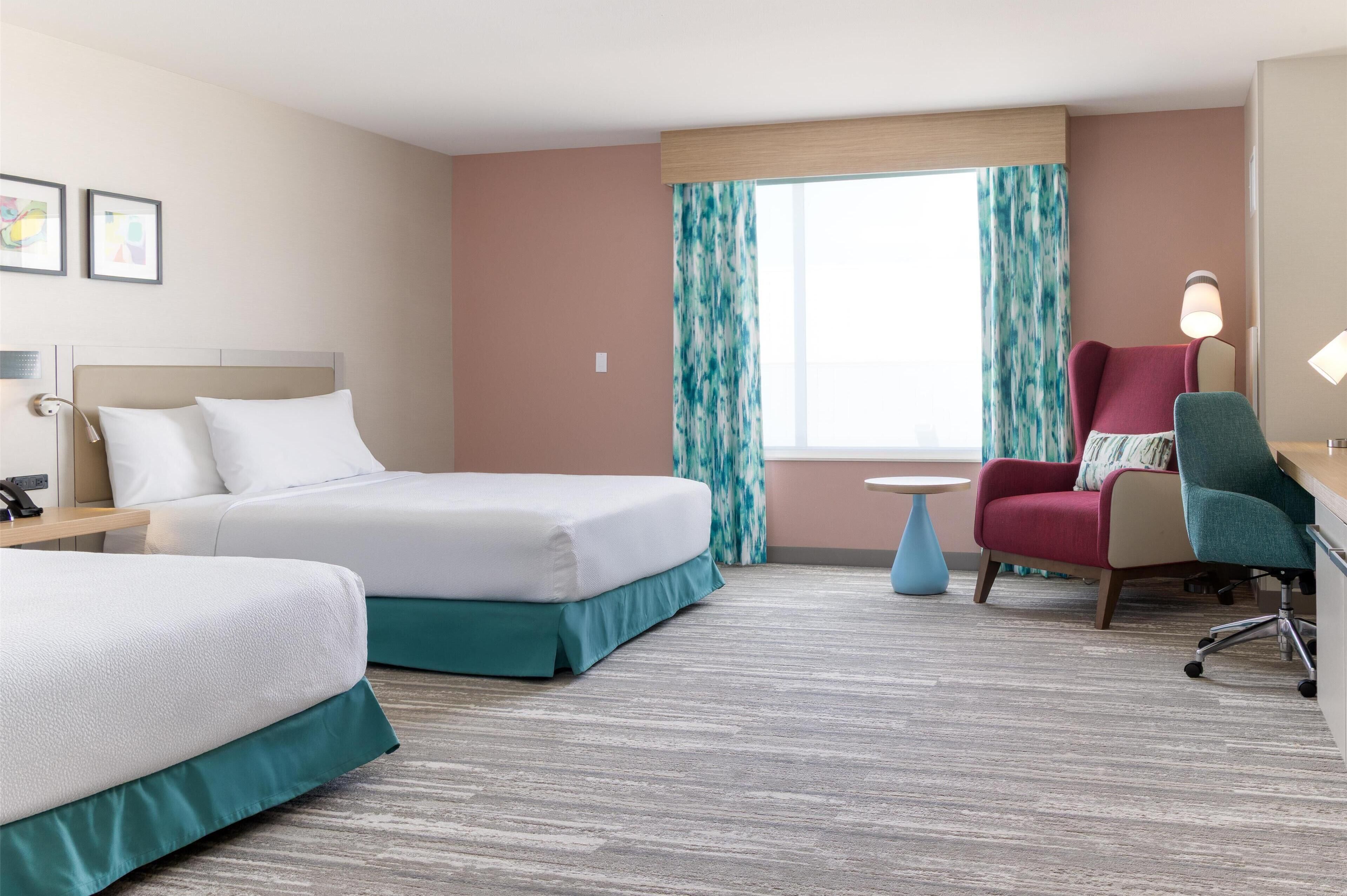 A hotel room with two beds, a desk, a chair, a lamp, and a window with curtains.