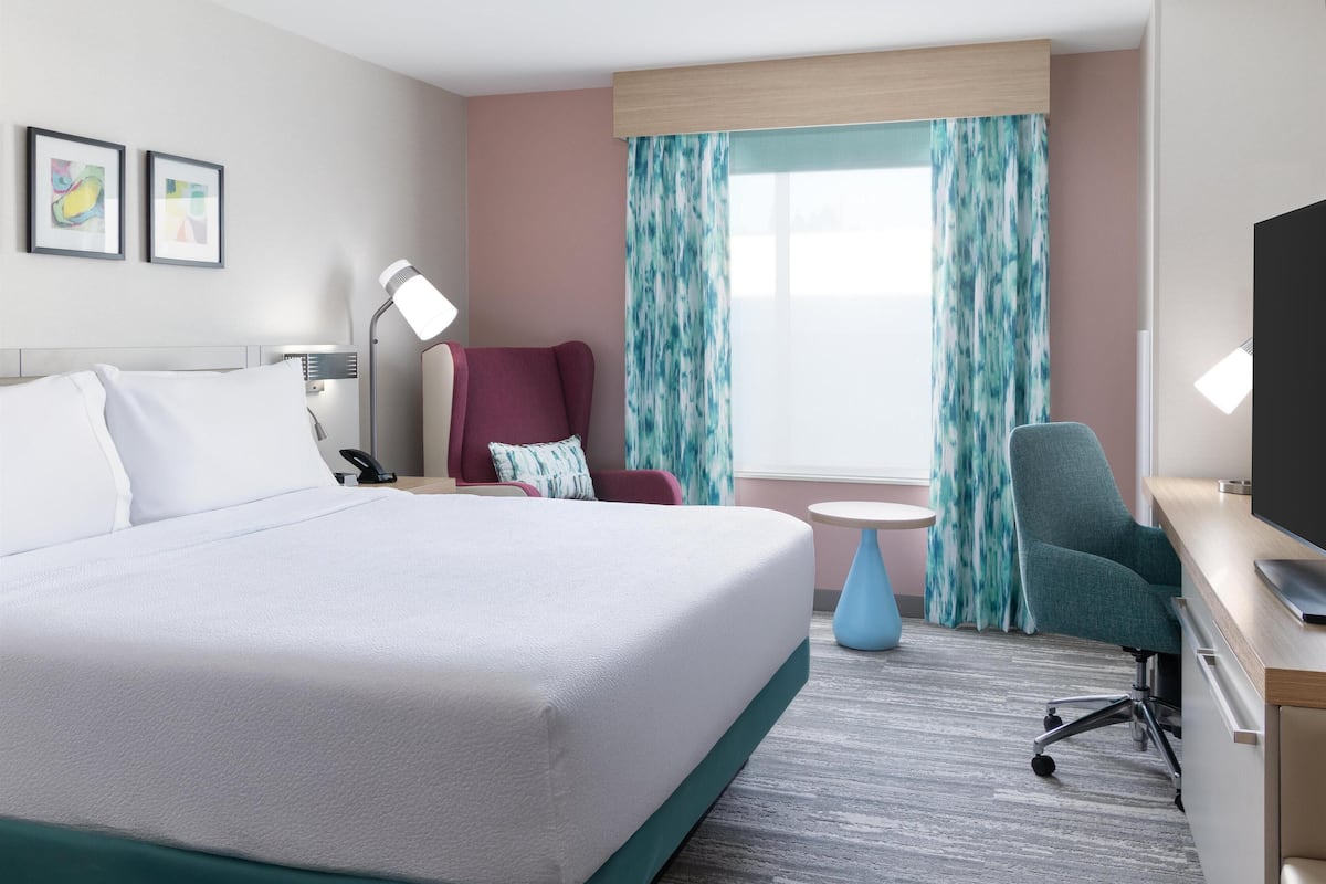 Habitación de hotel con una cama grande, un escritorio con televisor, una silla, una lámpara y una ventana con cortinas.