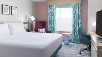 A hotel room with a large bed, a desk with a TV, a chair, a lamp, and a window with curtains.