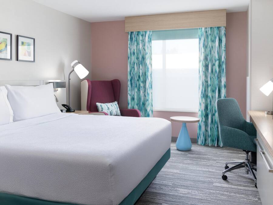 A hotel room with a large bed, a desk with a TV, a chair, a lamp, and a window with curtains.
