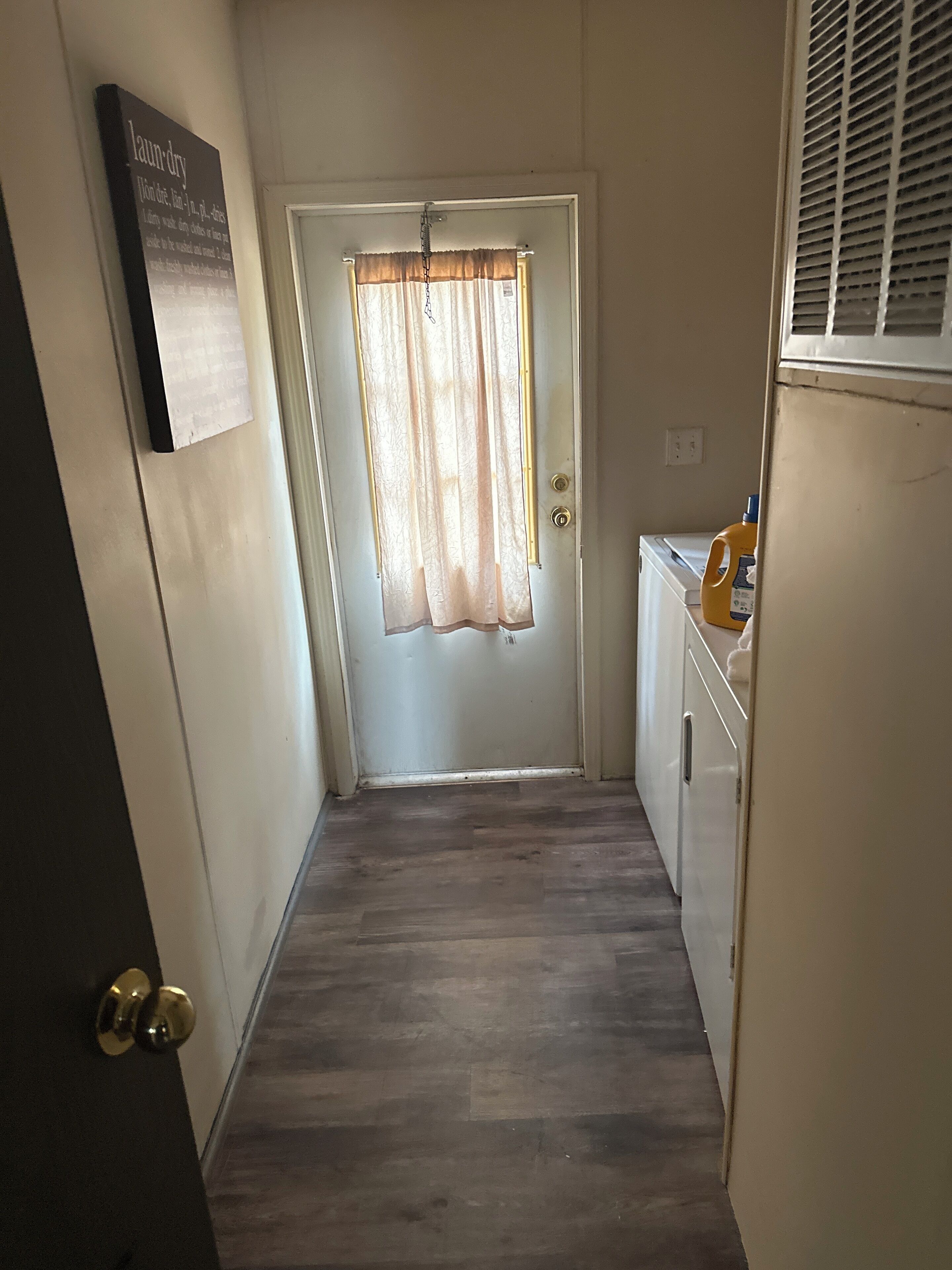 A narrow hallway with a door at the end, a small cabinet with a bottle of detergent, and a sign on the wall.