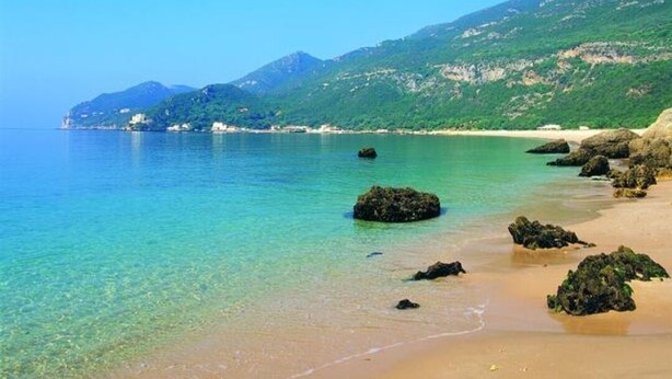 Uma praia com águas azul-turquesa cristalinas, areal arenoso e afloramentos rochosos.