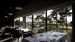 Um restaurante com mesas preparadas para refeições, grandes janelas que oferecem vista para árvores e um campo de golfe.