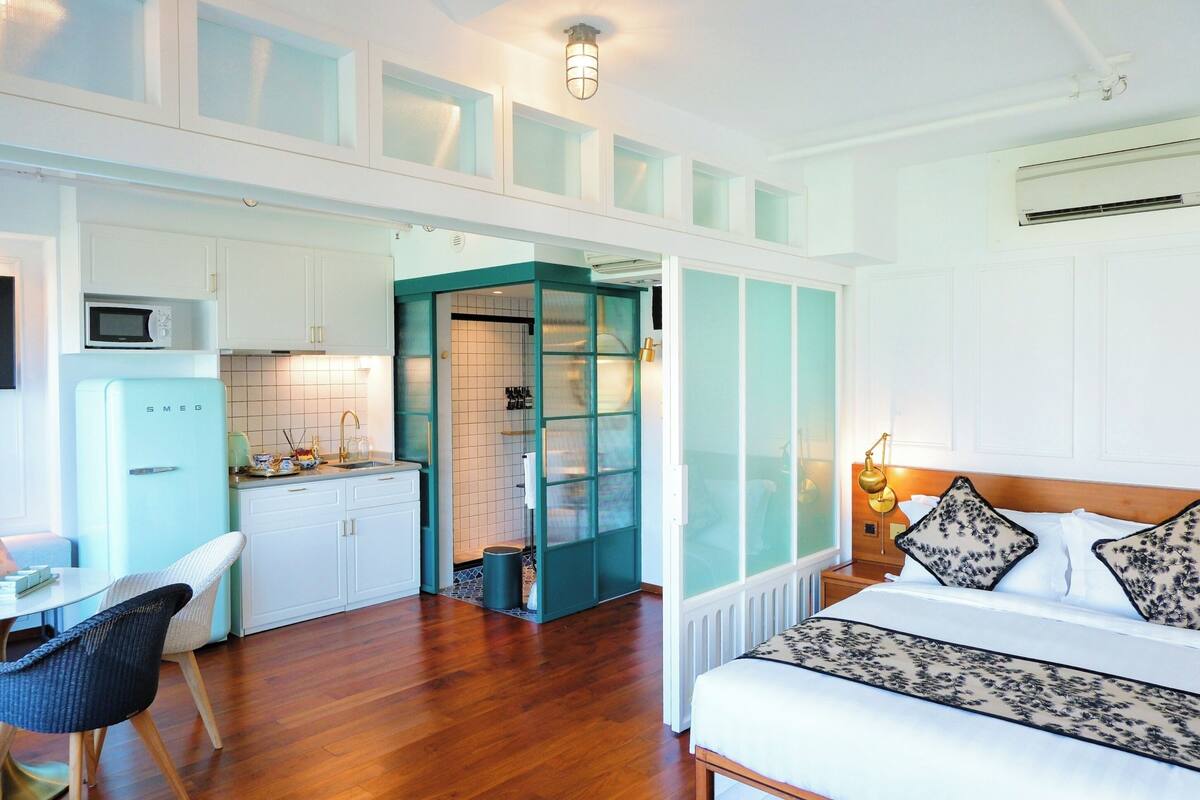 A modern hotel room with a wooden floor, a bed with patterned pillows, a white desk with a chair, a kitchenette with a microwave, and a bathroom with a glass door.