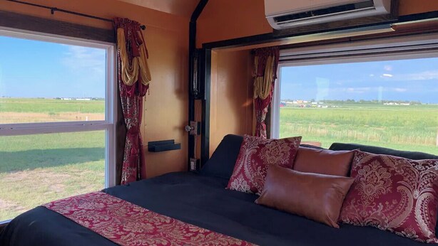A bedroom with a large bed, two windows with curtains, an air conditioning unit, and a view of a green field.