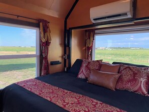 A bedroom with a large bed, two windows with curtains, an air conditioning unit, and a view of a green field.