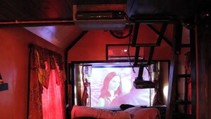 A room with a bed, a television screen displaying a person, and a red curtain.