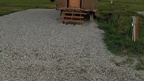 A small, single-story wooden structure with a door and a window, situated on a gravel path with a grassy field in the background.