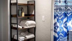 Un baño con un perchero con toallas dobladas, una cortina de ducha con un estampado geométrico y un interruptor de luz en la pared.
