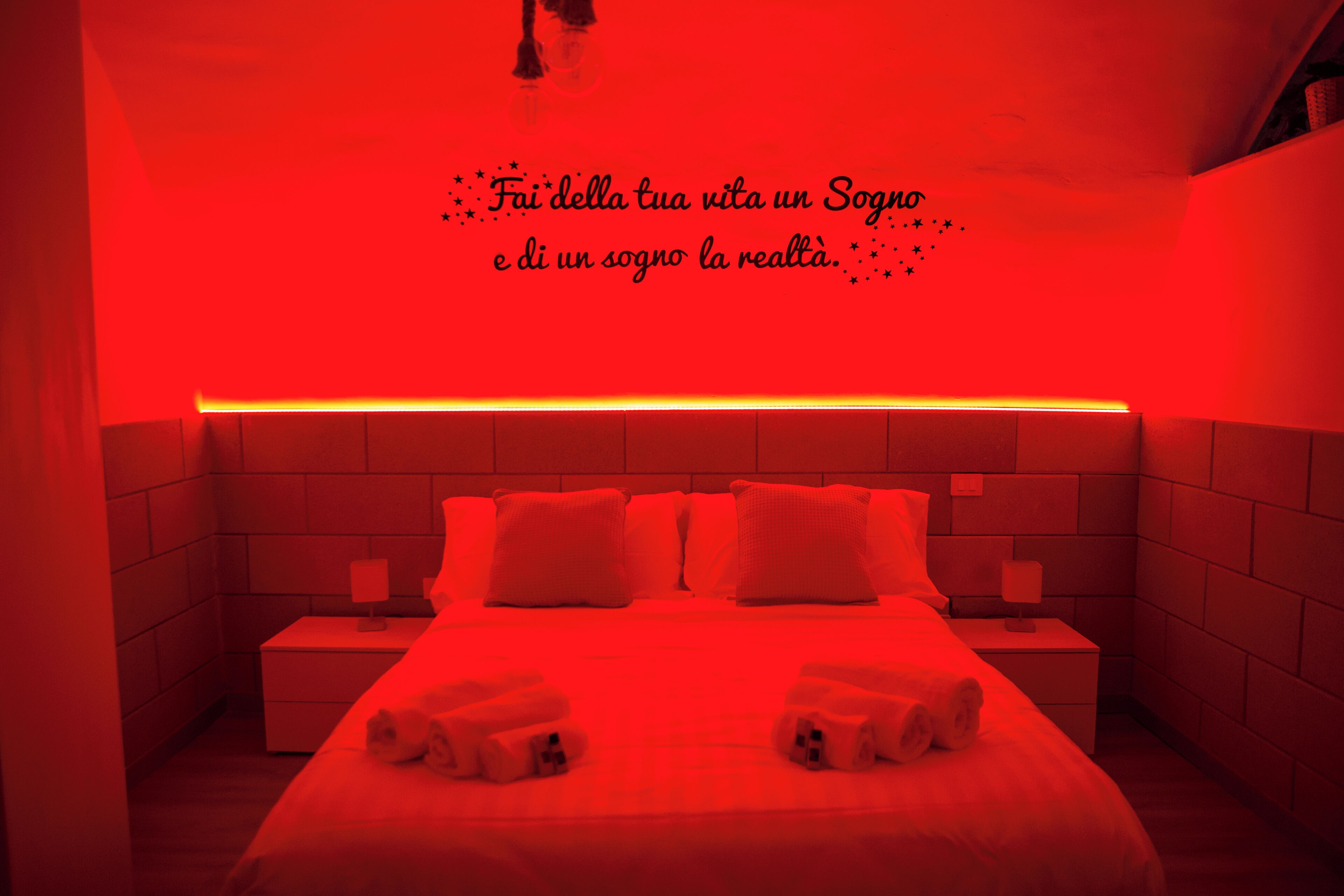 Une chambre d’hôtel éclairée de lumière rouge, avec un lit, des oreillers et une table de chevet sur laquelle se trouve une lampe.