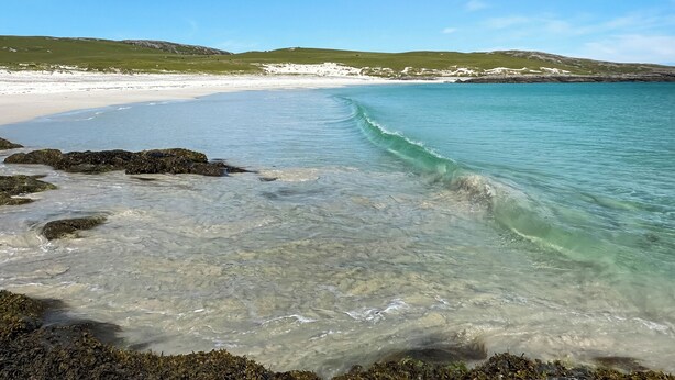 Una playa con agua cristalina, costa rocosa y colinas al fondo.