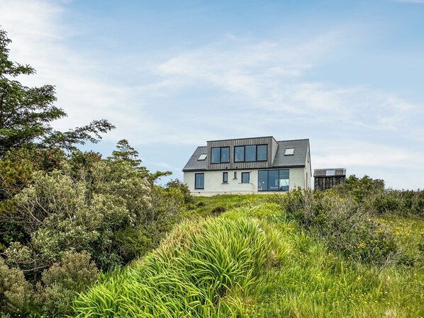 A modern two-story house with large windows and a sloped roof, surrounded by dense greenery.