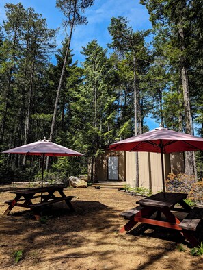 Dos mesas de picnic con sombrillas rojas, una terraza de madera y una pequeña construcción de madera en un área boscosa.