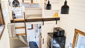 A compact kitchen with a stove, sink, and modern appliances. A loft bedroom with a bed and a fan is visible above the kitchen. - Cozy Tiny Home - Farm Stay - Near Qualicum Beach  (Qualicum Beach)