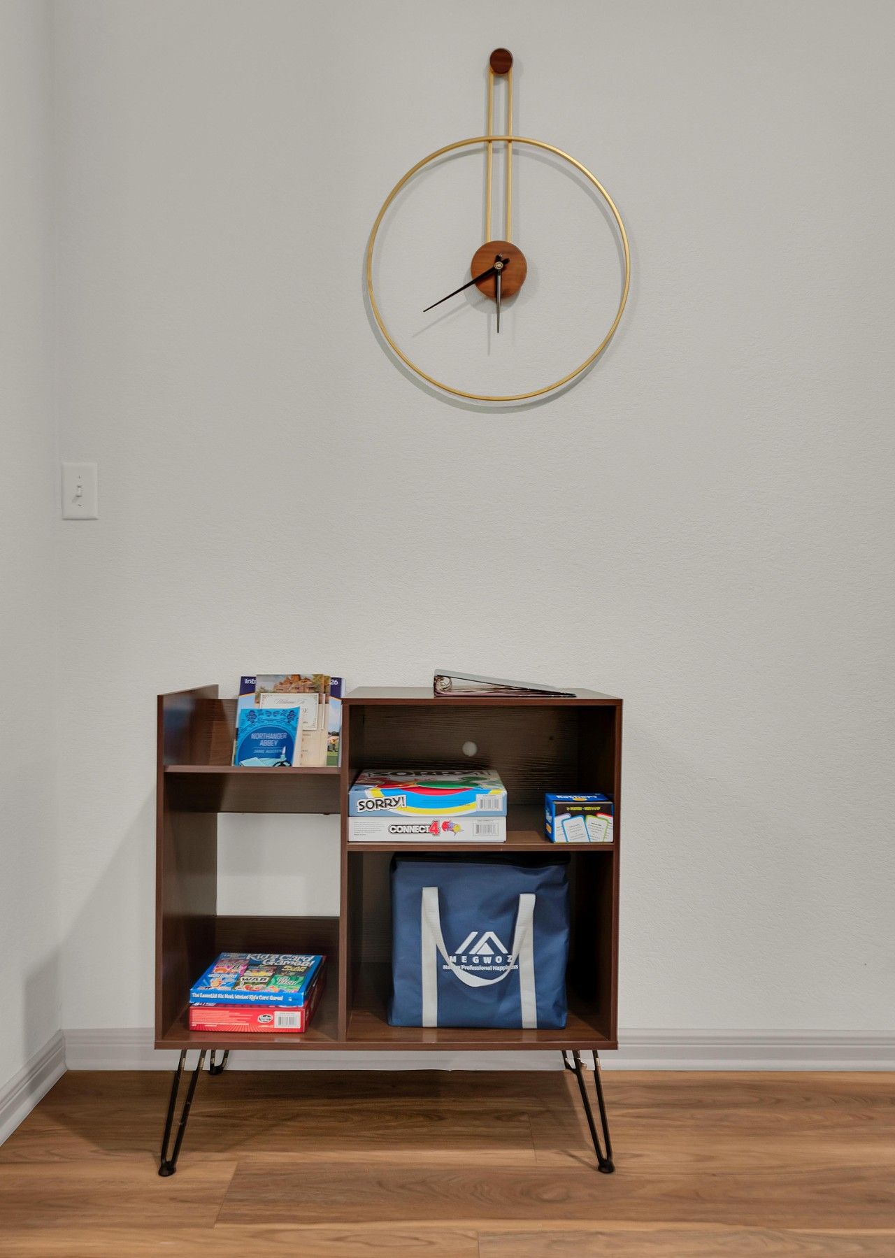 A room with a wooden floor, a wall-mounted clock, and a bookshelf with various items including books and a blue bag.