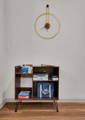 A room with a wooden floor, a wall-mounted clock, and a bookshelf with various items including books and a blue bag.