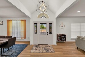 A spacious living room with a dining area, a large window with blinds, a modern chandelier, and a bookshelf.