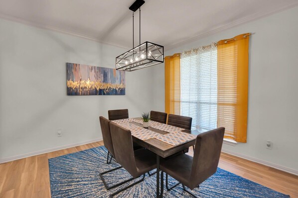 Un coin repas comprenant une table rectangulaire, six chaises, un luminaire suspendu moderne, un tapis à motifs bleus et une grande peinture abstraite accrochée au mur.
