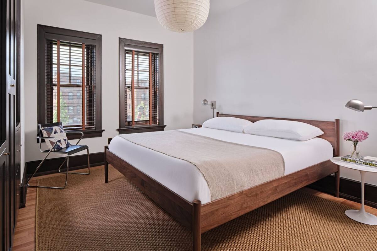 Um quarto com cama de madeira, duas janelas com persianas, uma cadeira, mesa de cabeceira com abajur e luminária pendente.