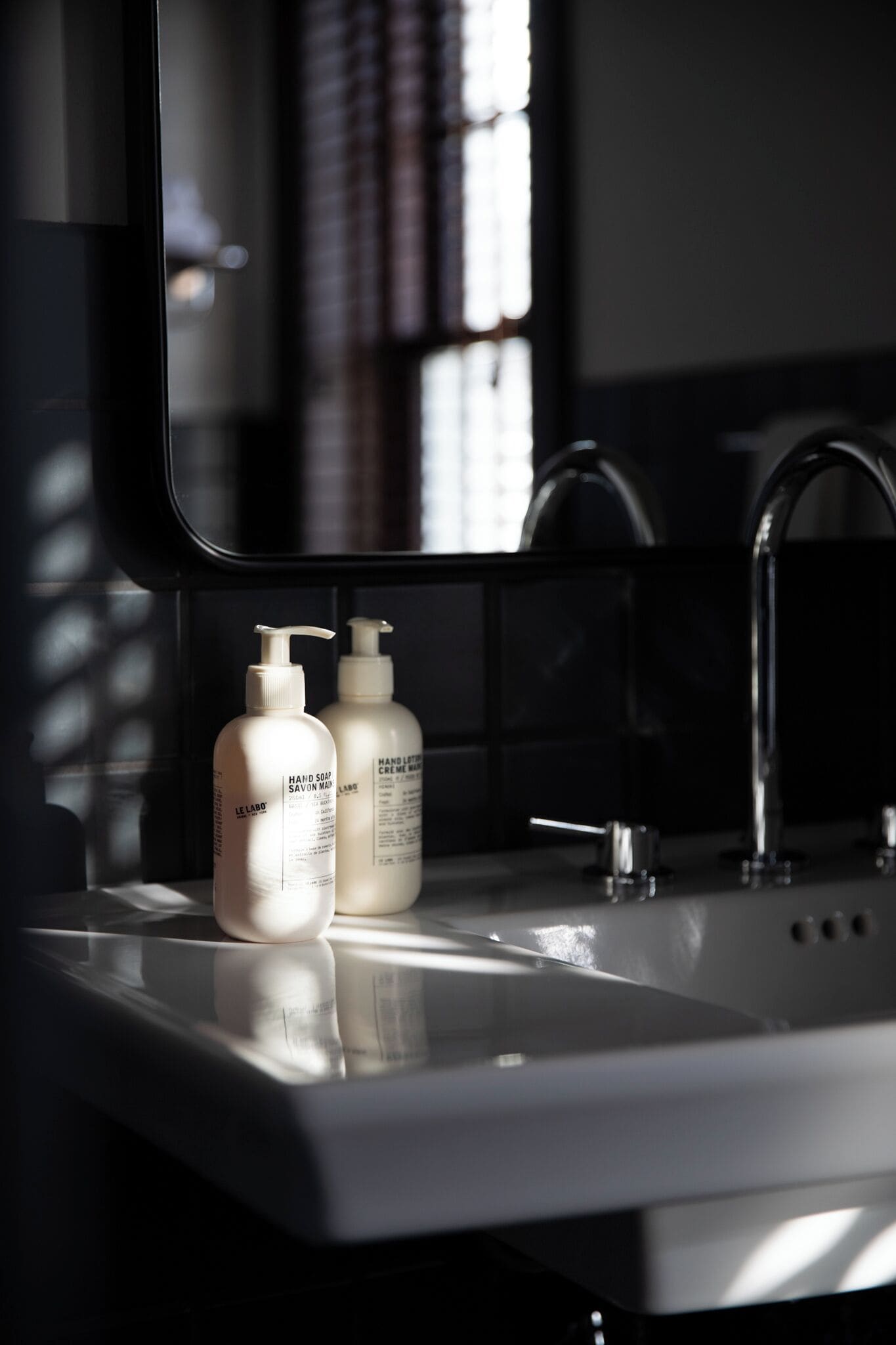 A bathroom with a white sink, two bottles of hand soap, and a mirror reflecting a window with blinds.