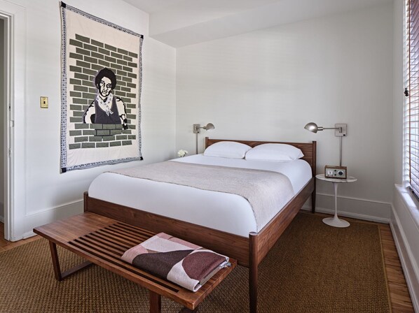Un dormitorio ordenado con una cama de madera, un banco, un cuadro en la pared y una mesita con un radio.