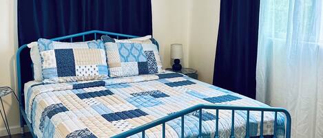 Un dormitorio con una cama, ventilador de techo, mesita de noche y ventana con cortinas.