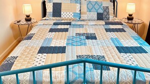 Ein Bett mit blauem Metallgestell, einem dunkelblauen Vorhang und einer Steudecke mit verschiedenen Mustern. Zwei Nachttische mit Lampen stehen daneben.