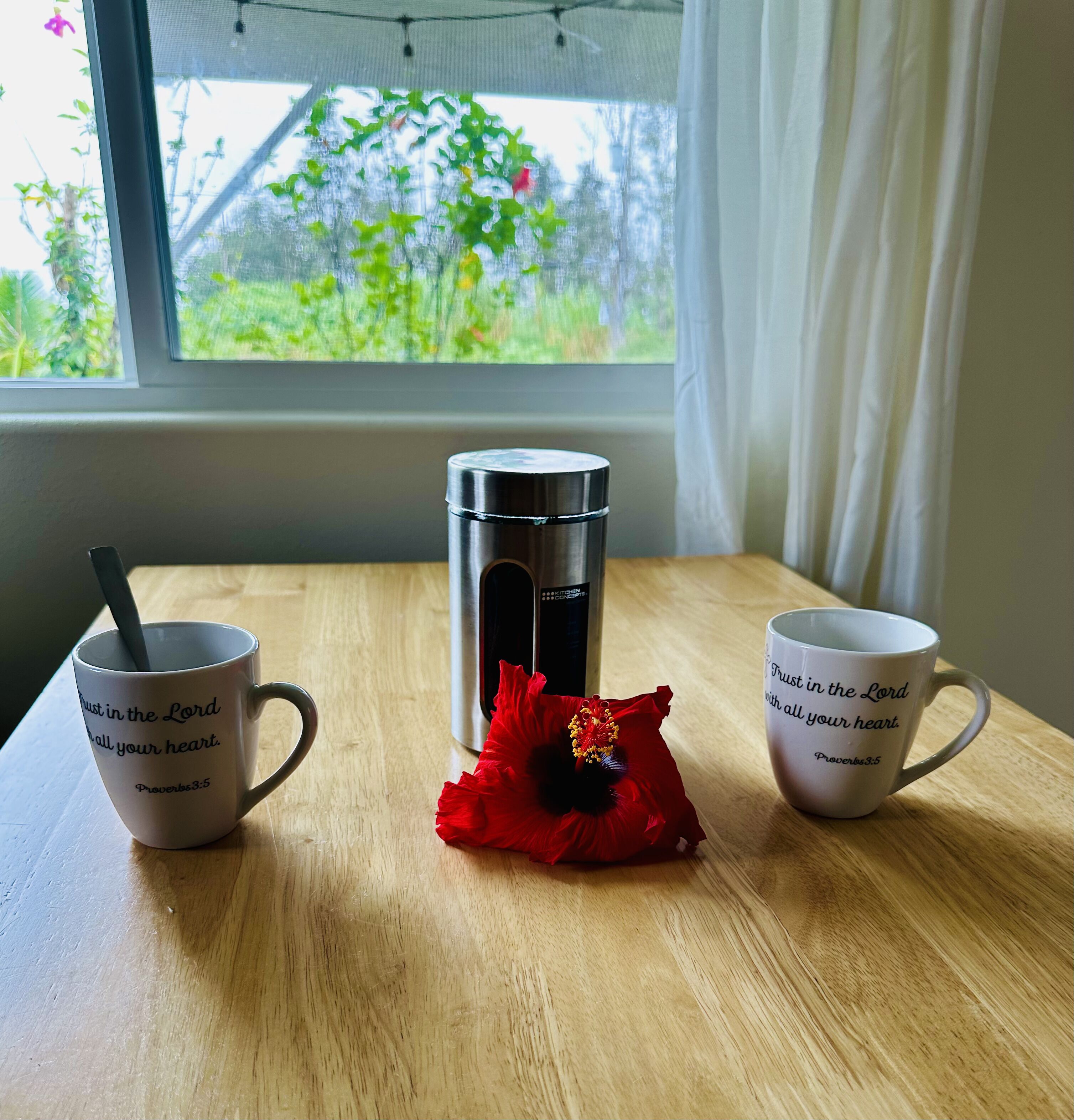 A wooden table with two white mugs, one with a black handle and the other with a white handle, and a stainless steel thermos.