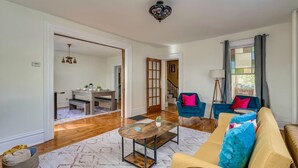 A spacious living room with a wooden coffee table, a beige sofa with blue cushions, and a blue armchair. There is a staircase leading to another level, and a dining area with a wooden table and benches. - Pool Table & More Spacious Family Fun (Canton)